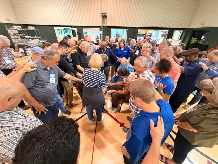 Large group prayer circle with hands on shoulders in a community hall